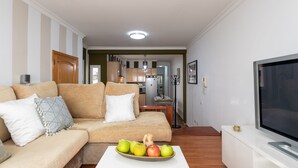 Ein Wohnzimmer mit einer Couch, einem Couchtisch mit Früchten, einem Fernseher und einer Küche im Hintergrund.