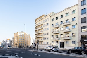 Un edificio de varios pisos con balcones, una calle con coches y un cielo despejado.