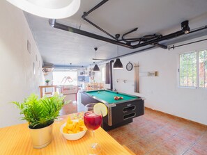 A pool table with a drink, a bowl of chips, and a potted plant in a room with a kitchen area, a clock, and a window.