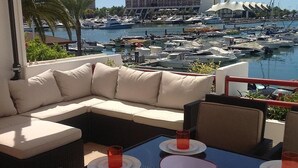 A balcony with a sofa and dining table overlooking a marina with boats and a hotel in the background. - Luxury 3 Bedroomed Apartment- Stunning Views, Direct Marina Access & Shared Pool (Vilamoura)