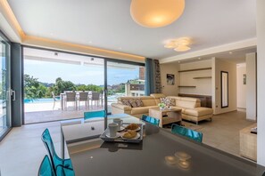 Un salon moderne avec un canapé, une table à manger où sont disposés des aliments pour le petit-déjeuner, et une vue sur une piscine et la verdure environnante.