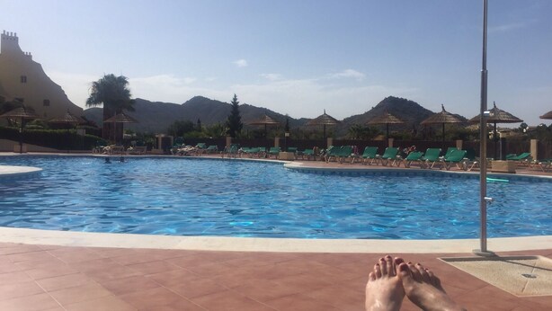 Una piscina con sillas de descanso y sombrillas, rodeada de montañas y un edificio con torre.