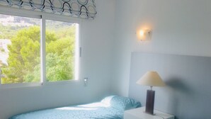 Un dormitorio con dos camas individuales, una lámpara de noche, una ventana con vistas a zonas verdes, y una lámpara fijada en la pared.