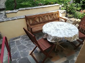 Eine Terrasse im Freien mit einem runden Tisch, bedeckt mit einer blumigen Tischdecke, umgeben von Holzstühlen und -bänken.