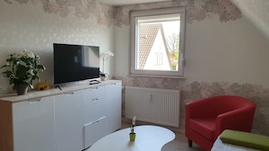 A compact living space with a TV on a cabinet, a red chair, a small table, and a window with a view of buildings outside.