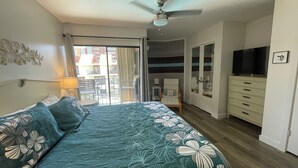 A bedroom with a bed, a ceiling fan, a TV, and a balcony with a view of buildings.
