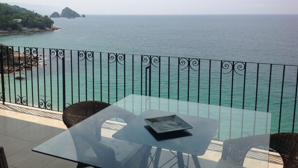 Ein Balkon mit Glastisch und Korbstühlen, der auf ein türkisfarbenes Meer und entfernte Land blickt.