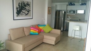 Ein Wohnzimmer mit einem beigefarbenen Ecksofa, bunten Kissen, einem gerahmten Kunstwerk und einer Küchenzeile mit Kühlschrank und Mikrowelle.
