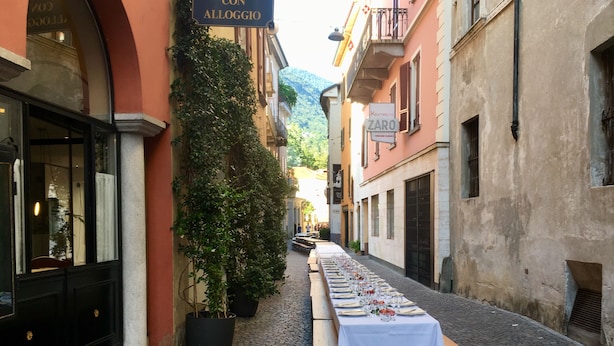 Una estrecha calle adoquinada con una larga mesa dispuesta para una comida, bordeada por edificios con letreros que señalan un restaurante y un hotel.