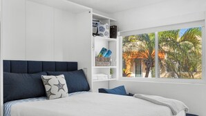 Un dormitorio con una cama, una mesita de noche, estante empotrado con toallas y ventana con vista a palmeras.