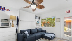 Una sala moderna con un sofá seccional, un ventilador de techo, un microondas, una lámpara, un ventanal con vistas a palmeras y una señal de salida.