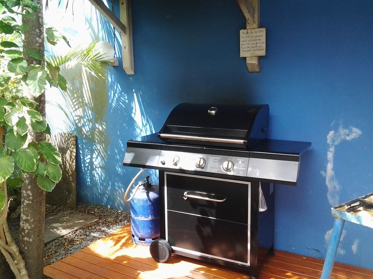 A barbecue grill with a gas tank, a drawer, and a handle, positioned on a wooden deck against a blue wall.