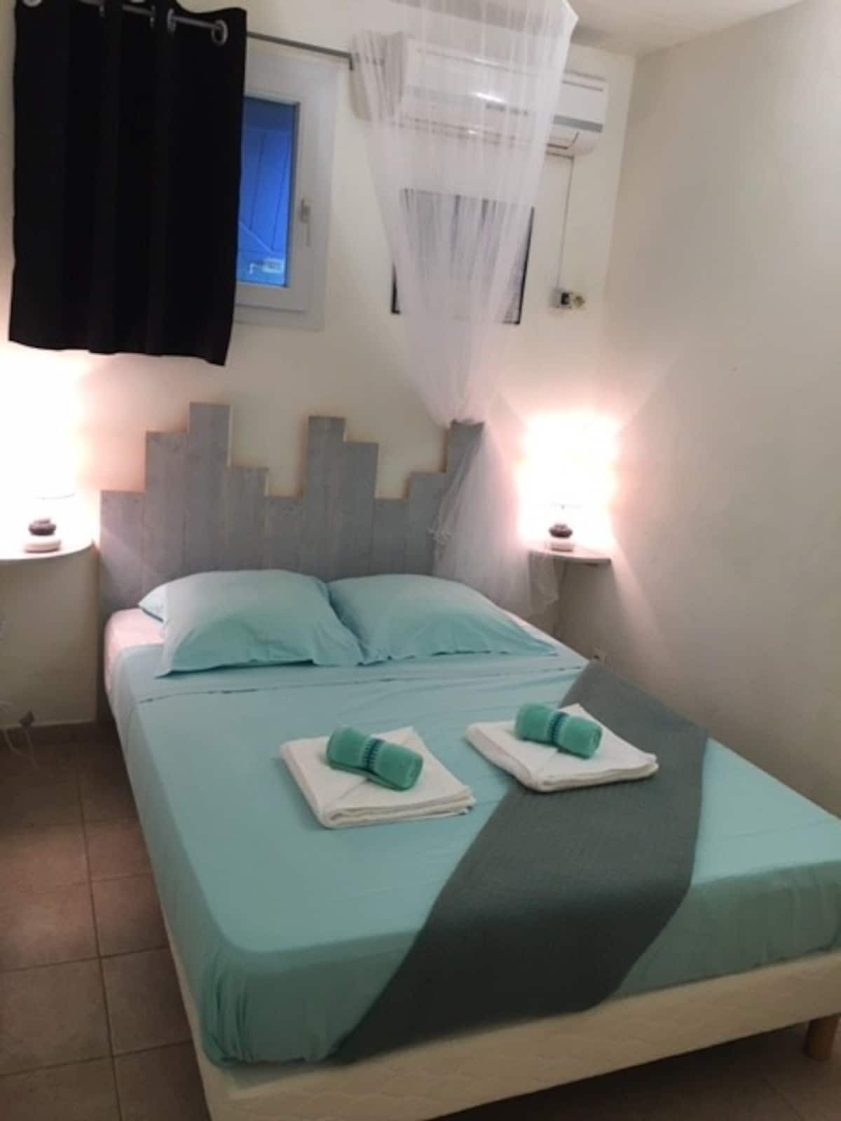 A single bed with a grey headboard, two pillows, and two folded towels. There are two bedside lamps, a window with a curtain, and an air conditioning unit on the wall.