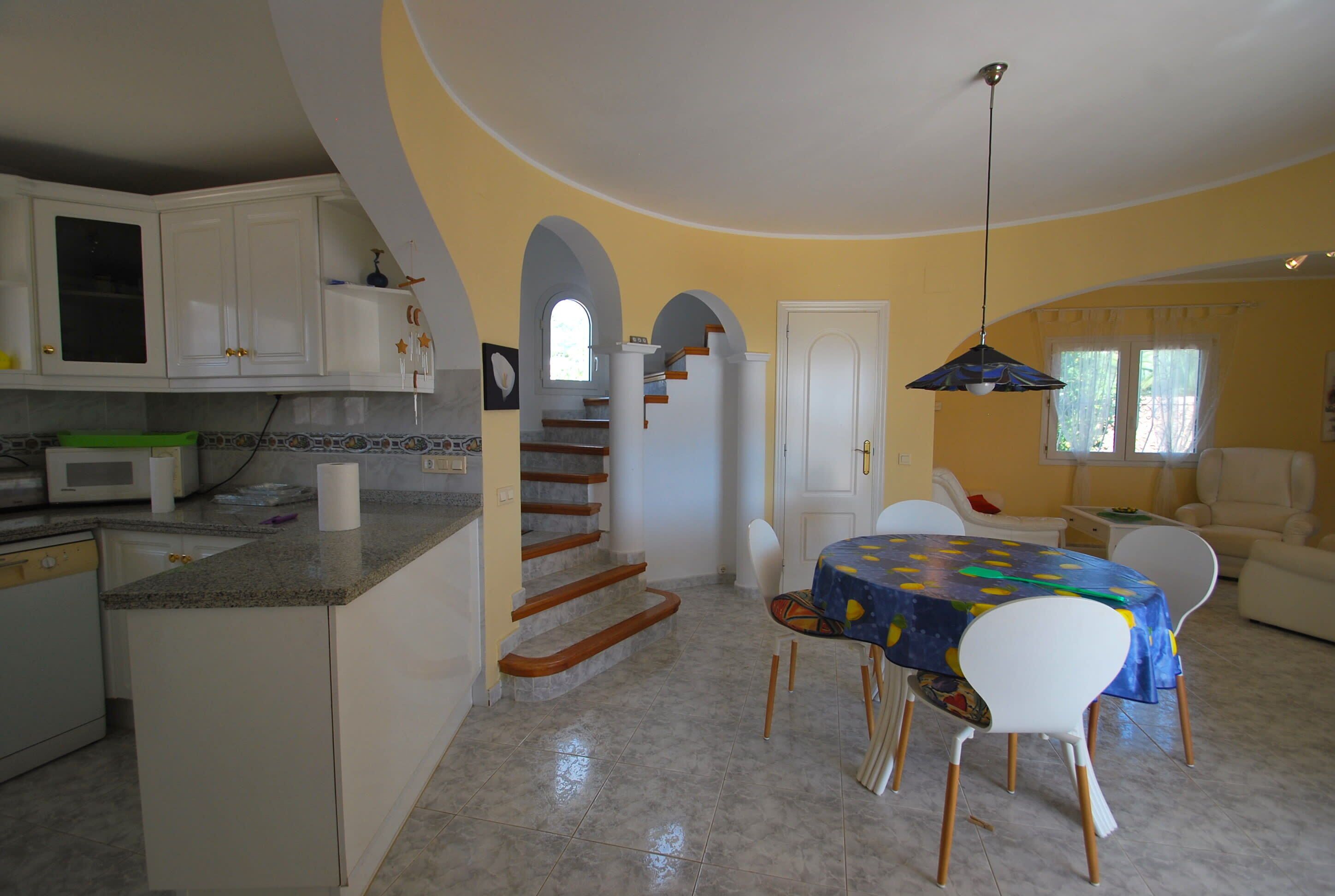 Une cuisine avec des meubles blancs, un coin repas comprenant une table et des chaises, et un escalier menant à un autre niveau.