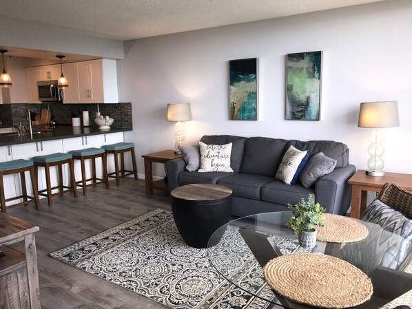 A modern living room with a grey sofa, a coffee table, and a kitchen area with bar stools. - Beautifully Furnished, New Renovation, A/C, Mtn/Ocean View, beach/golf/hiking! (Waianae)