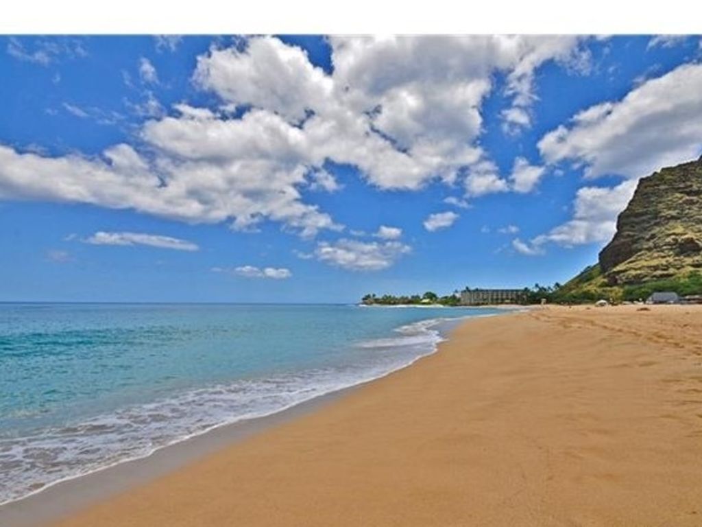 A beach with clear blue water, a sandy shore, and a building on a hill in the background.