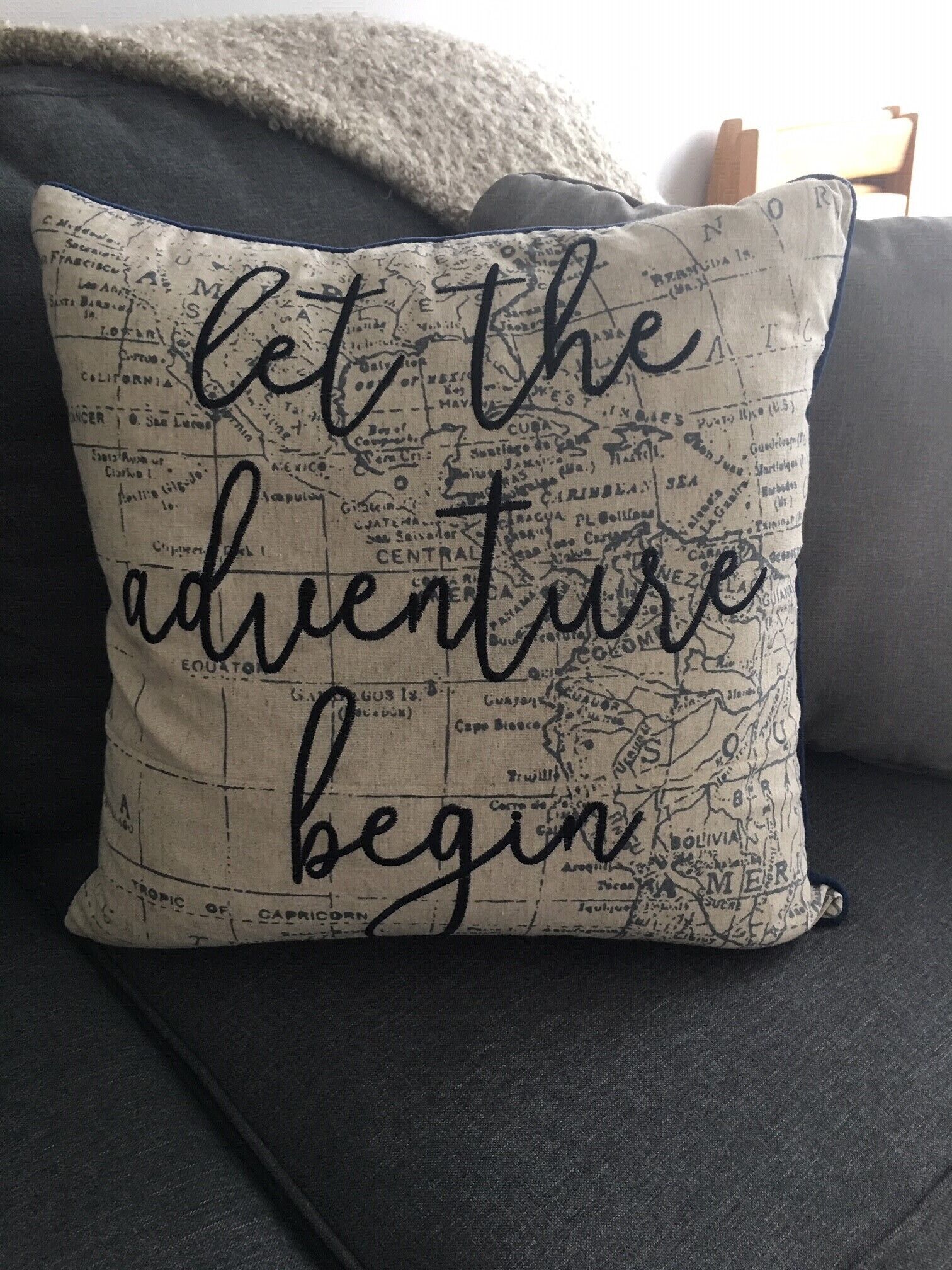 Un cuscino con una mappa e la scritta "let the adventure begin" su un divano scuro.