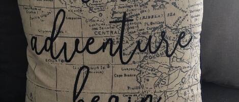 Un cuscino con una mappa e la scritta "let the adventure begin" su un divano scuro.