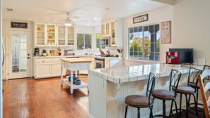 A kitchen with white cabinets, a central island, bar stools, and a large window. - Waterfall Spa + Sunset Views | Atascadero Wine Country | Summer Place (Templeton)