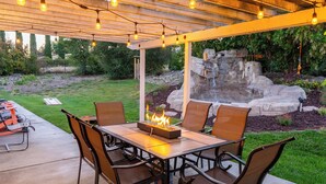 An outdoor patio with a table and chairs, a fire feature, and a stone waterfall backdrop. - Waterfall Spa + Sunset Views | Atascadero Wine Country | Summer Place (Templeton)