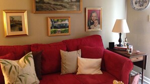 Ein rotes Sofa mit Kissen, ein gerahmtes Landschaftsbild, ein gerahmtes Foto und eine Lampe auf einem Beistelltisch.