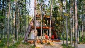 Una cabaña en un bosque con una terraza de madera, dos sillas y un pequeño porche.
