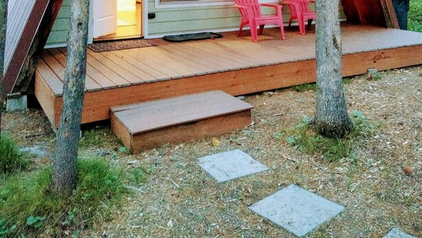 Una cabaña con terraza de madera, dos sillas Adirondack rosa y un amplio ventanal con vistas al bosque.