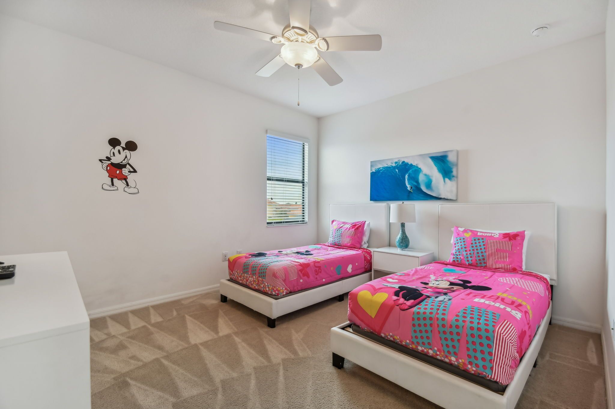 Een slaapkamer met twee bedden, een plafondventilator, een raam met jaloezieën en een Mickey Mouse sticker op de muur.