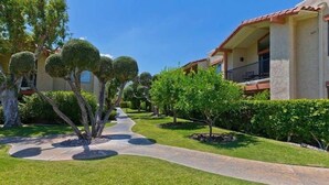 Ein gepflegter Garten mit einem gepflasterten Weg, ordentlich geschnittenen Hecken und einem Gebäude mit einem Ziegeldach und Balkonen.