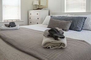 Un lit bien fait, recouvert d’une couette grise et blanche, une commode blanche avec des tiroirs et une lampe posée sur le dessus.