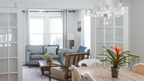 Ein geräumiges Wohnzimmer mit einem großen Esstisch, weißen Stühlen und einem Kronleuchter. Im Hintergrund befindet sich eine Couch und ein Couchtisch mit einer Lampe.