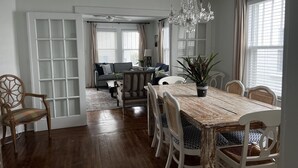 Ein Essbereich mit Holztisch und weißen Stühlen, einer Kandelaber und Blick auf ein Wohnzimmer mit einer Couch und Vorhängen.