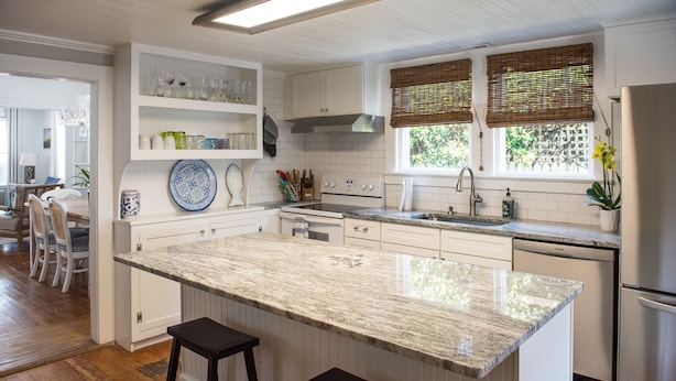 Eine moderne Küche mit weißen Schränken, einer Granitarbeitsplatte und einem eingebauten Backofen. Zwei Barhocker und ein Fenster mit Jalousien sind vorhanden.