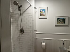 Une salle de bain avec des murs recouverts de carreaux blancs, une douche avec un pommeau de douche amovible, deux tableaux encadrés accrochés au mur et un porte-rouleau de papier toilette.