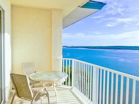A balcony with a glass table and chairs, overlooking a body of water with a distant shoreline.
