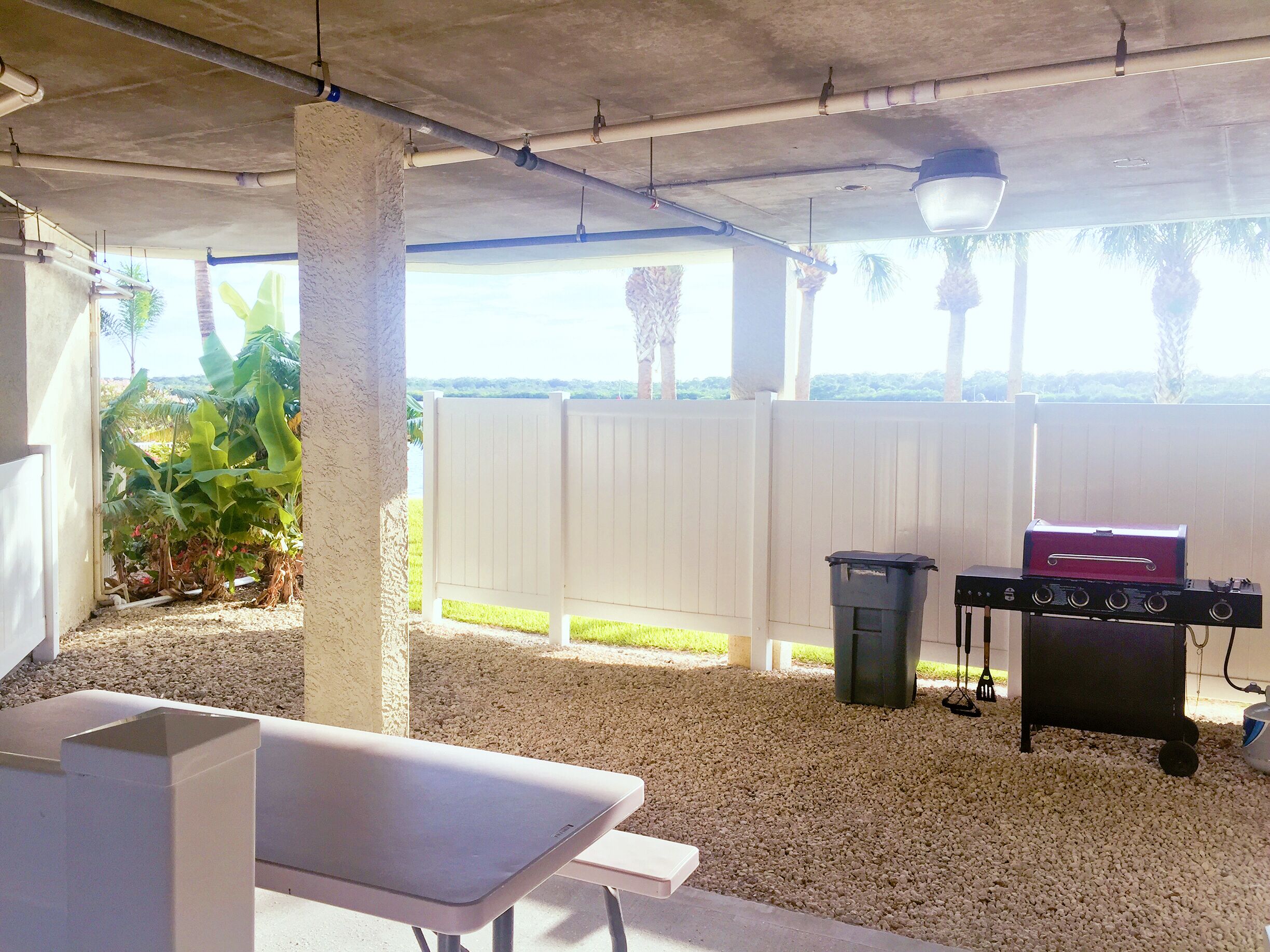 An outdoor patio area with a barbecue grill, a white fence, and a view of palm trees.