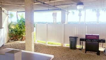 An outdoor patio area with a barbecue grill, a white fence, and a view of palm trees.