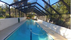 A long, rectangular swimming pool with clear blue water, surrounded by a covered area with a brick wall and a metal roof structure. - Getaway New Smyrna Beach, Sun, Pool, Shopping. (New Smyrna Beach)