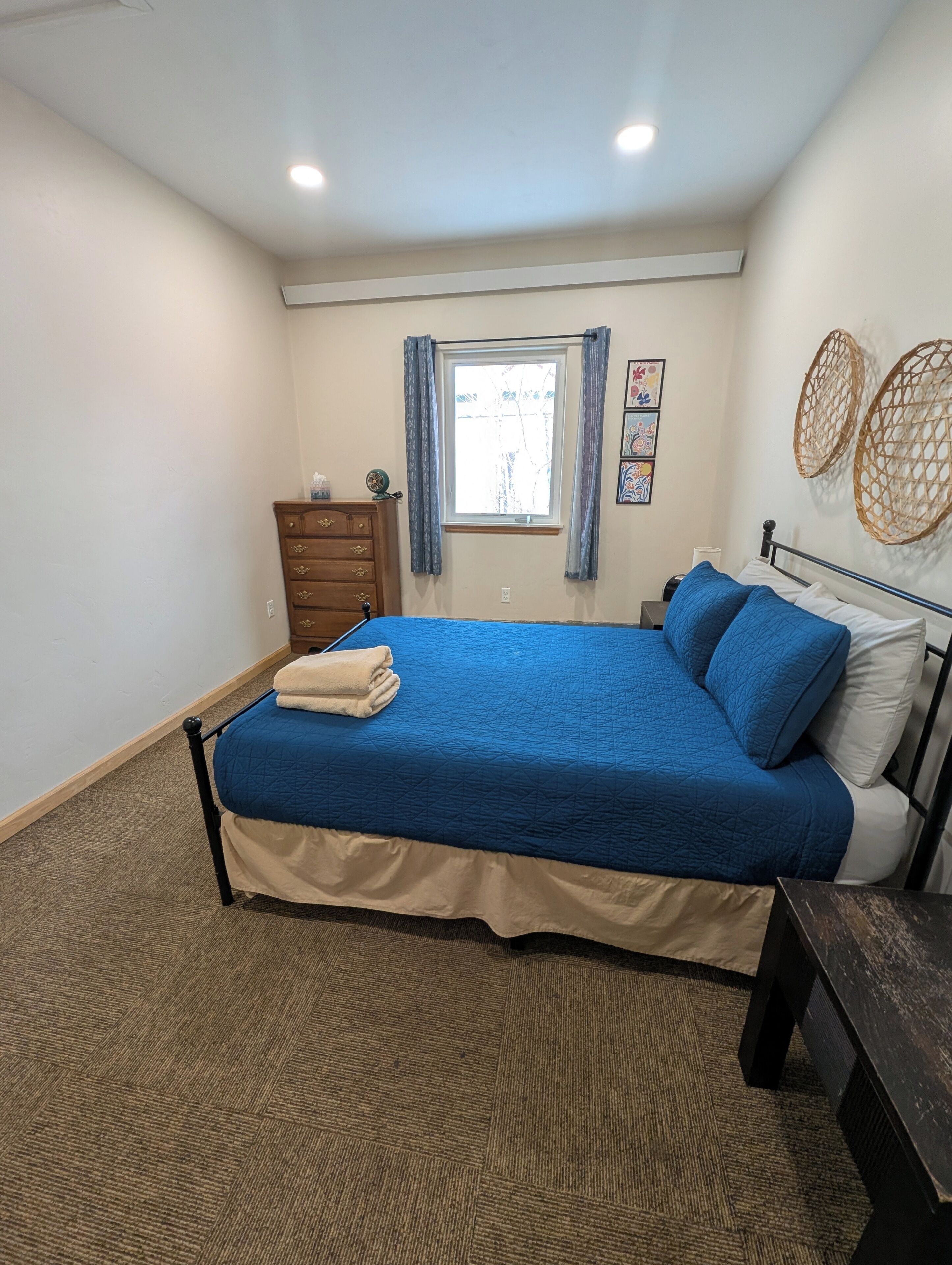 A bedroom with a bed, a dresser, a nightstand, a window with curtains, and a wall-mounted decorative item.