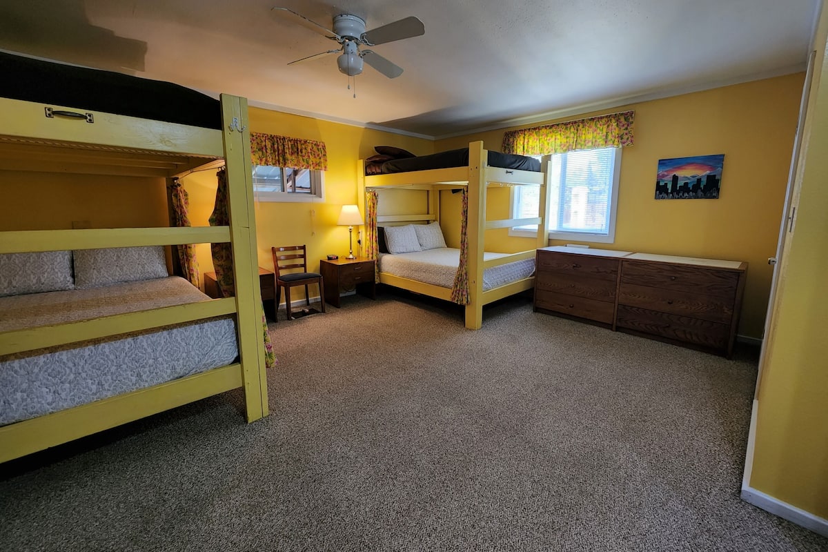 A dormitory room with bunk beds, a desk, a chair, a lamp, a dresser, and a ceiling fan.