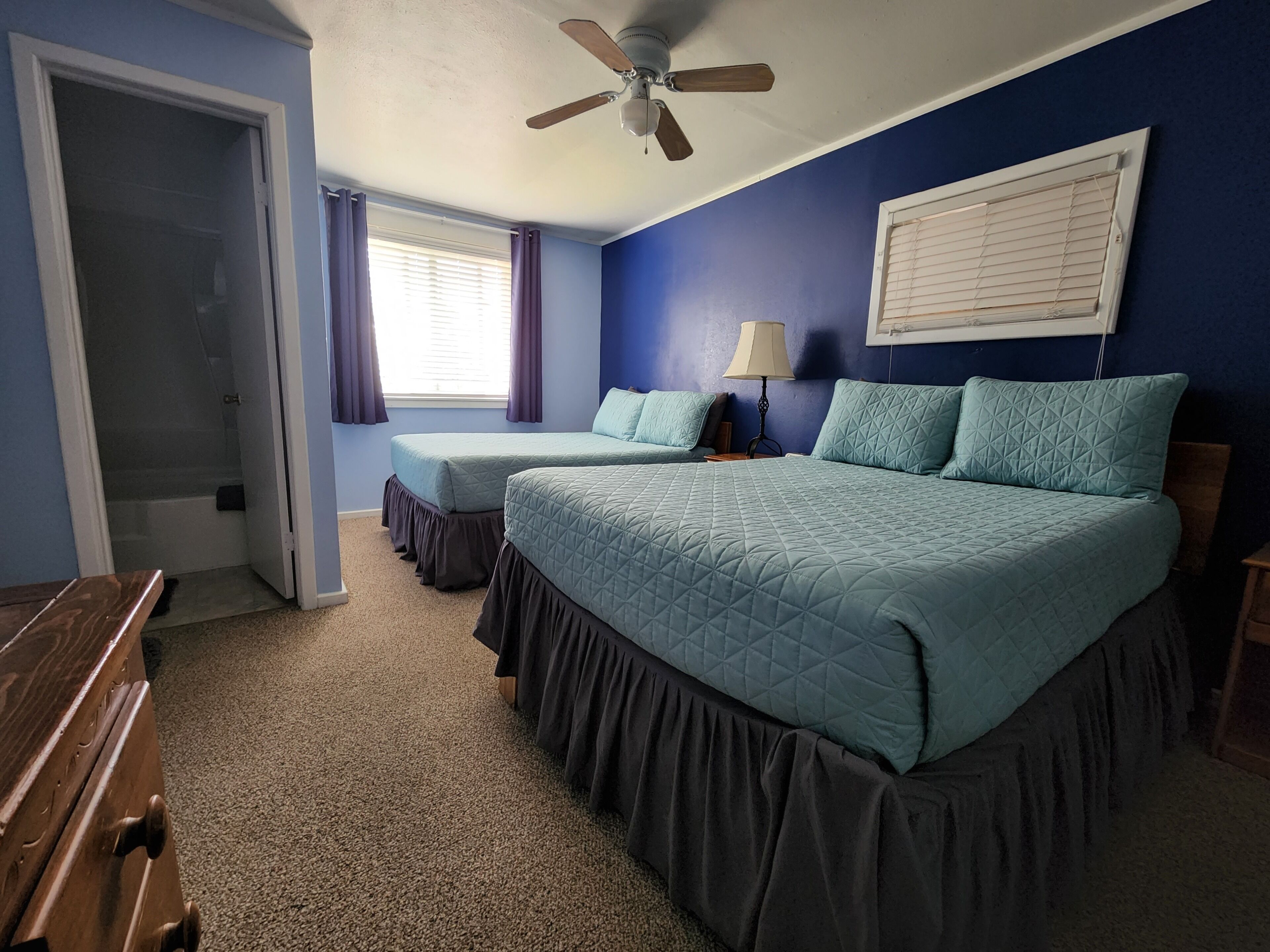 Une chambre avec deux lits, un ventilateur de plafond, une fenêtre avec des rideaux et une porte donnant sur une salle de bain.