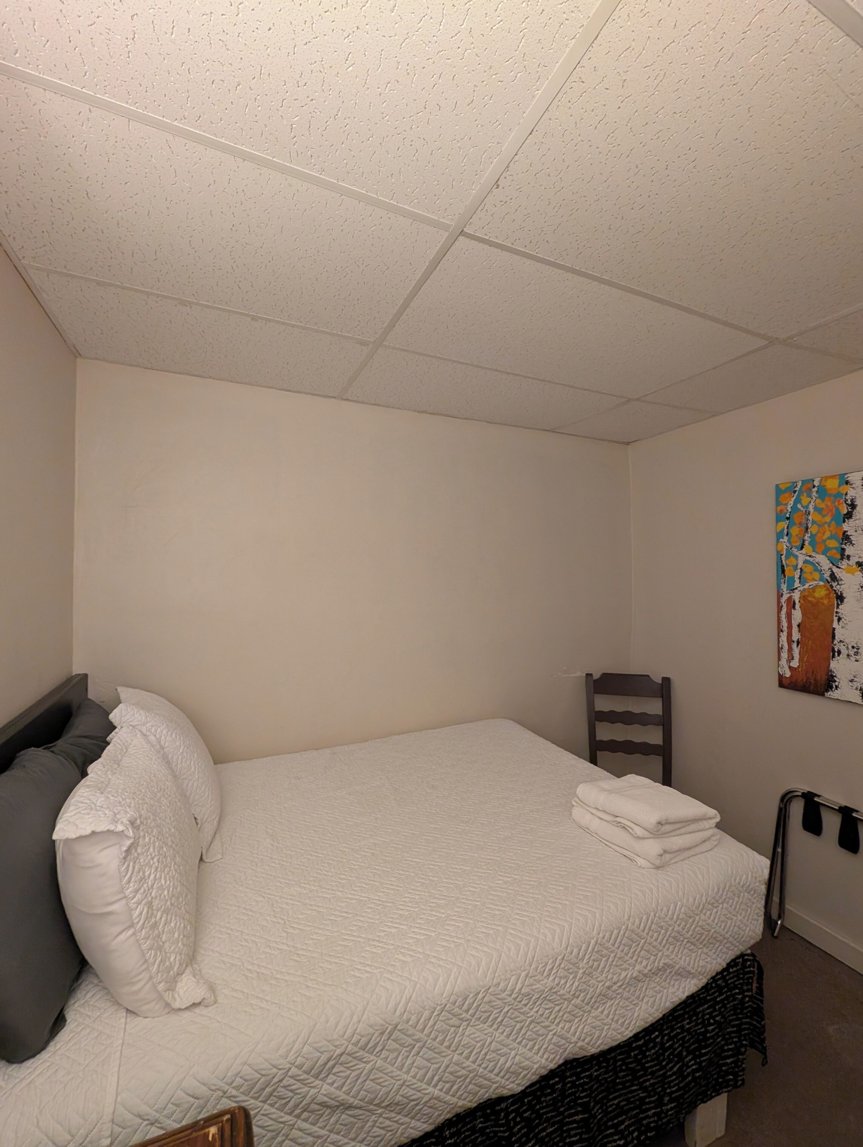 A single bed with a white quilt, a gray pillow, and a wooden chair in a room with plain walls and a ceiling with square tiles.