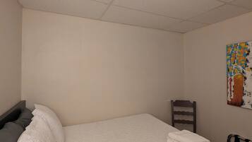 A single bed with a white quilt, a gray pillow, and a wooden chair in a room with plain walls and a ceiling with square tiles.