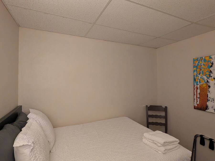 A single bed with a white quilt, a gray pillow, and a wooden chair in a room with plain walls and a ceiling with square tiles.