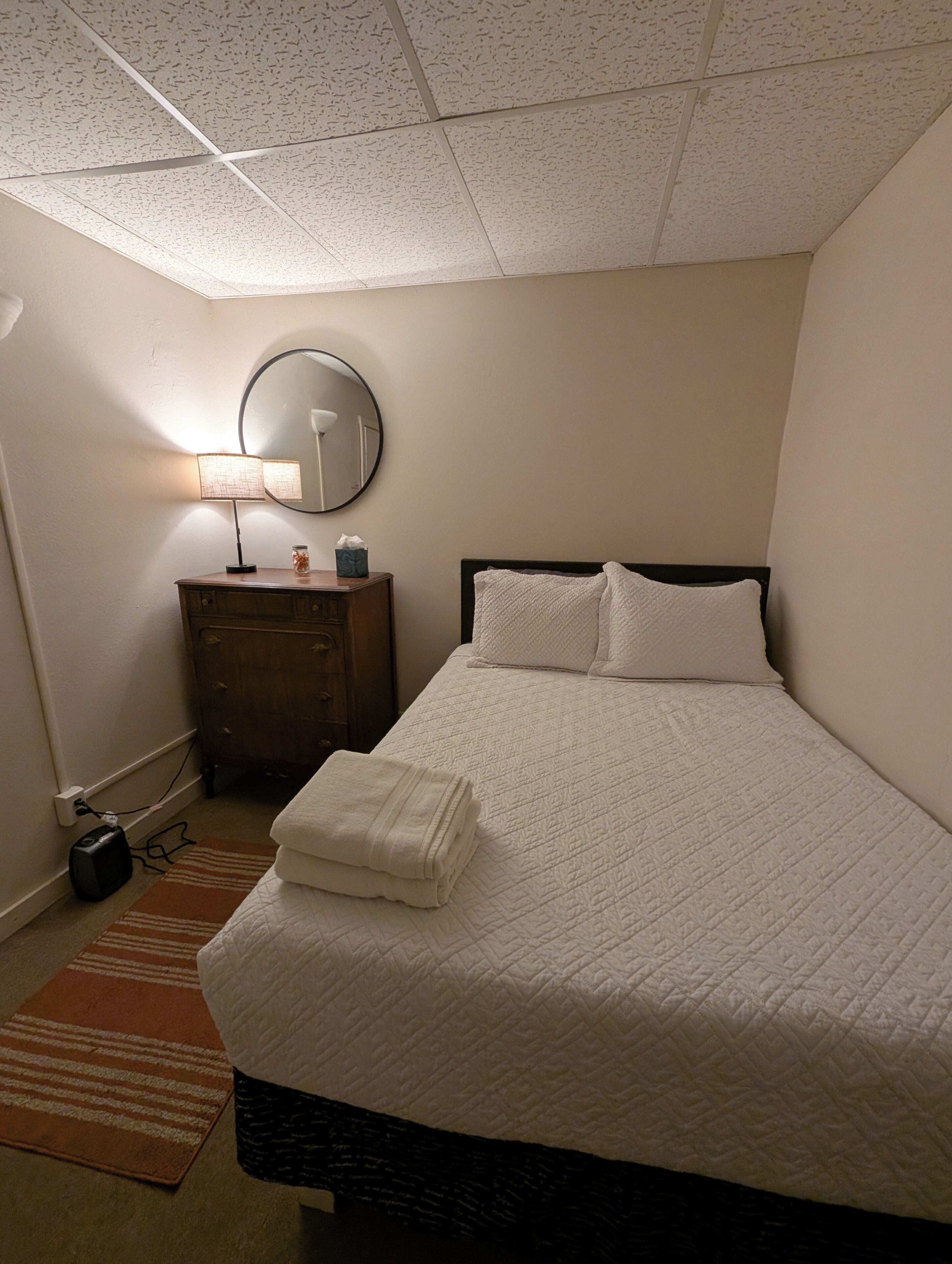 A neatly made bed with a white quilt, a bedside lamp, a mirror, and a wooden dresser.