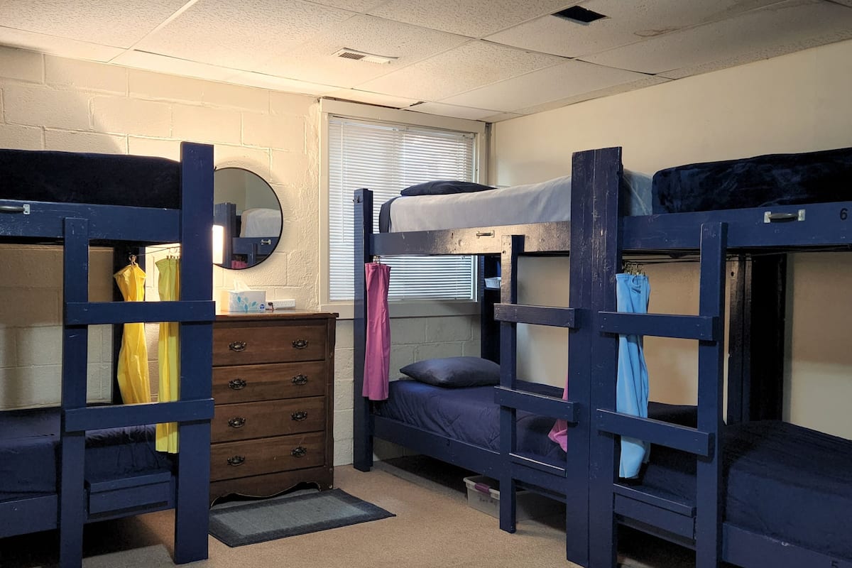 A dormitory room with bunk beds, a dresser, a mirror, and a window with blinds.