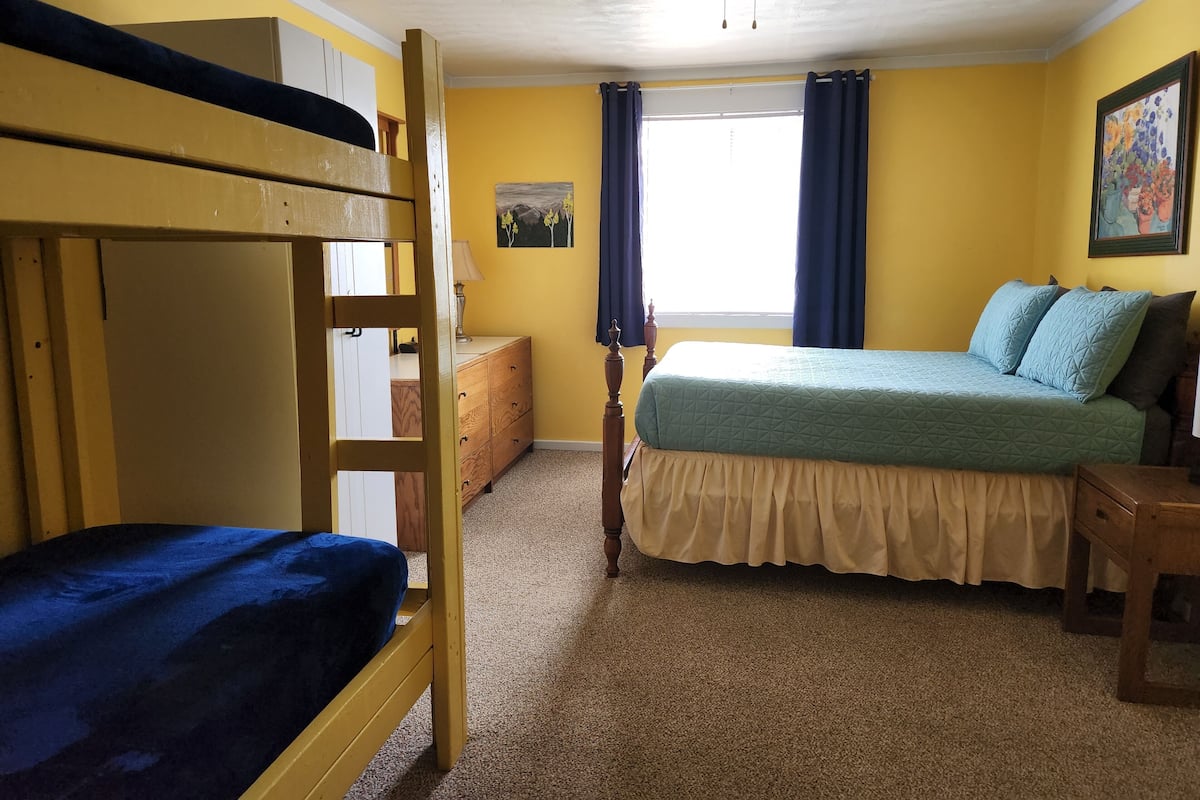 A room with a bunk bed, a single bed with a blue quilt, a wooden dresser, a lamp, and a ceiling fan.