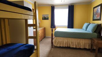 Um quarto com uma cama beliche, uma cama de solteiro com colcha azul, uma cômoda de madeira, um abajur e um ventilador de teto.