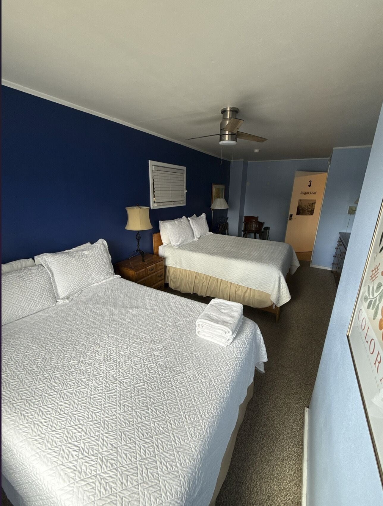 A hotel room with two beds, a ceiling fan, a nightstand, a lamp, and a door labeled 'Sugar Loft'.