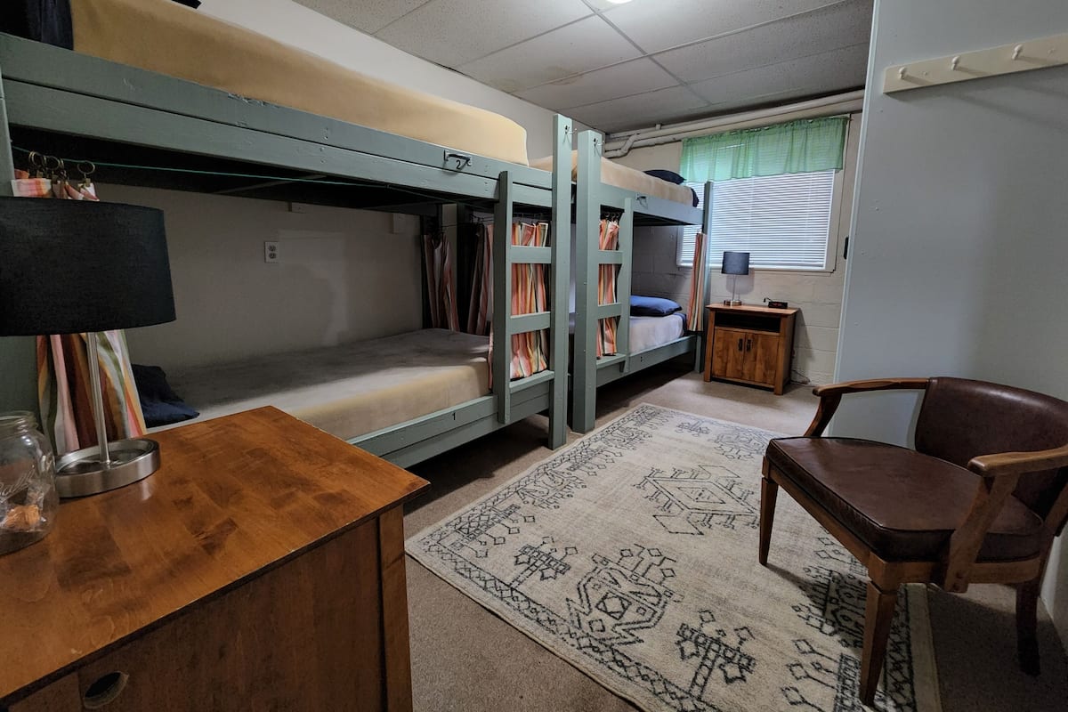 A dormitory room with bunk beds, a desk, a chair, a small cabinet, and a patterned rug.